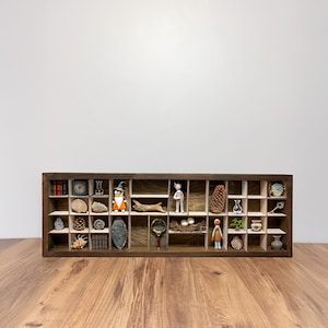 May include: A wooden display shelf with multiple compartments, each containing a unique miniature object. Items include a clock, shells, a witch figurine, a magnifying glass, a pinecone, and a small doll. The shelf is brown and sits on a wooden surface.