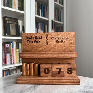 May include: A wooden book tracker display featuring a shelf with three small vertical book-shaped blocks and three larger square blocks displaying the number 075. The top panel is engraved with the text Books Read This Year and Christopher Smith.