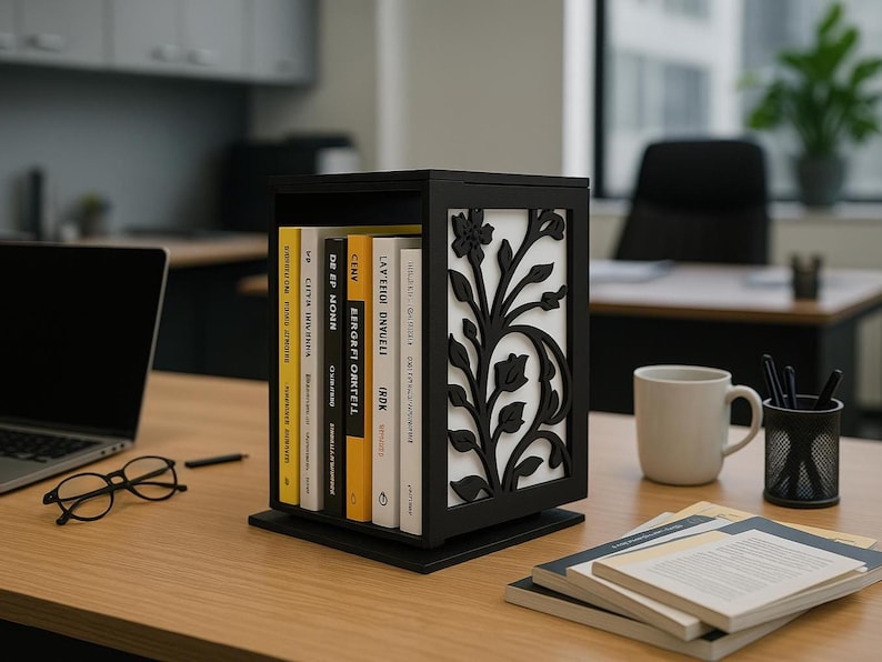 A black wooden book holder with floral cutout side panels and a white background stands on a wooden office desk. It holds seven upright books. Around it are a laptop, round eyeglasses, a white coffee mug, a black mesh pencil holder with pens, and stacked open notebooks. The background shows a modern office with another desk, black chairs, and a large window with a potted plant.