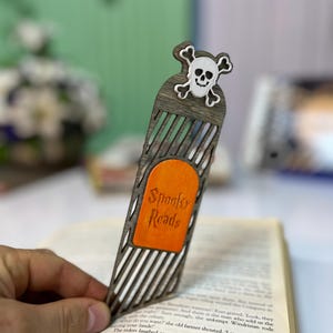 Peut inclure: Marque-page en bois avec un motif de tête de mort et d'os croisés. Le marque-page a une finition en grain de bois foncé avec une section rectangulaire orange avec l'inscription "Spooky Reads". Le marque-page est inséré dans un livre ouvert.