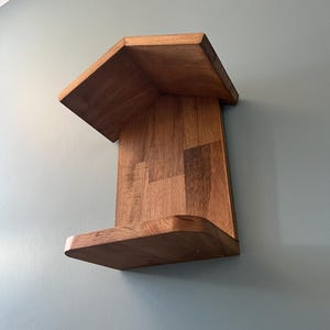 Puede incluir: Estantería esquinera de madera con acabado natural. La estantería tiene un diseño angular único con una balda superior e inferior. Montada en una pared azul claro, muestra su forma geométrica y veta de la madera.