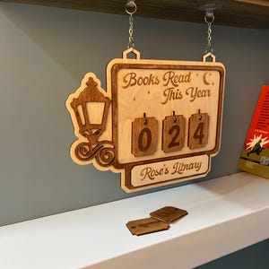 Puede incluir: Letrero de madera con el texto "Books Read This Year" y el número "024". Presenta un diseño decorativo de farola y el texto "Rose's Library". Incluye etiquetas numéricas de madera adicionales.