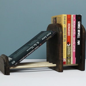 May include: Dark stained wooden bookends holding several books. The bookends have a unique design with a slanted section for displaying books. The books have colorful spines and titles like "1984" and "Remarque".