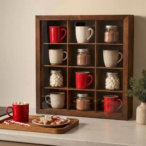Puede incluir: Una vitrina de madera con varias tazas, frascos y una pequeña planta de hoja perenne. Las tazas son rojas y beige. Los frascos contienen malvaviscos y cacao. Una taza roja con chocolate caliente y galletas.