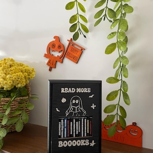 Könnte beinhalten: Ein schwarzes Bücherregal aus Holz mit den Worten "READ MORE" und "BOOOOKS" in Weiß. Das Bücherregal enthält Miniatur-Bücher zum Thema Halloween. Dekorative Holzausschnitte eines Kürbisses und ein Schild mit der Aufschrift "HAPPY HALLOWEEN" befinden sich an der Wand.