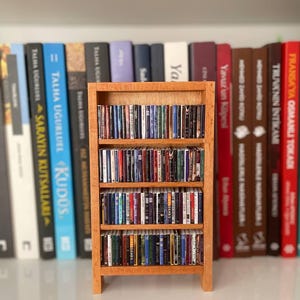 Peut inclure: Une petite bibliothèque en bois clair remplie de minuscules livres de différentes couleurs. La bibliothèque a trois étagères. En arrière-plan, une étagère contient des livres plus grands avec des titres en langue étrangère.