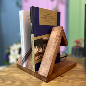 Puede incluir: Un sujetalibros de madera con un diseño triangular, que sostiene dos libros. Uno tiene una cubierta blanca y el otro una cubierta azul oscuro con letras doradas. El sujetalibros es de madera teñida y está sobre una superficie de madera.
