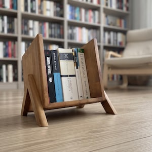 Op de afbeelding: Een lichtbruine houten boekenstandaard met schuine zijkanten en vier poten, met daarin vijf boeken, waaronder titels van George Orwell en Sandy Tolan. De standaard heeft een eenvoudig open ontwerp met een gladde afwerking en zichtbare boekruggen.