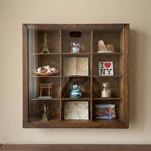 May include: A dark wood display case with a glass front, showcasing travel souvenirs. Items include a miniature Eiffel Tower, snow globes, a Colosseum replica, maps, shells, and an "I Love NY" sign. The case is mounted on a wall.