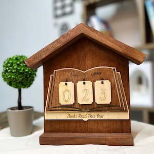 May include: A wooden house-shaped book counter with the text "Lisa's Library" and "Books Read This Year." The counter displays the number "023" on three small wooden tags. A small potted plant is to the left of the counter.