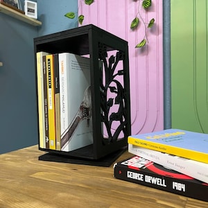 A black wooden book holder with floral cutout side panels is placed on a wooden surface. It holds five upright books inside, while three additional books, including George Orwell’s 1984, are stacked horizontally nearby. The background features a pastel green and purple wall with vertical grooves and hanging faux vines.