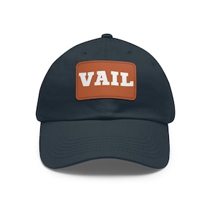 Op de afbeelding: Een marineblauwe baseballpet met een bruin leren patch waarop "VAIL" in witte letters staat.