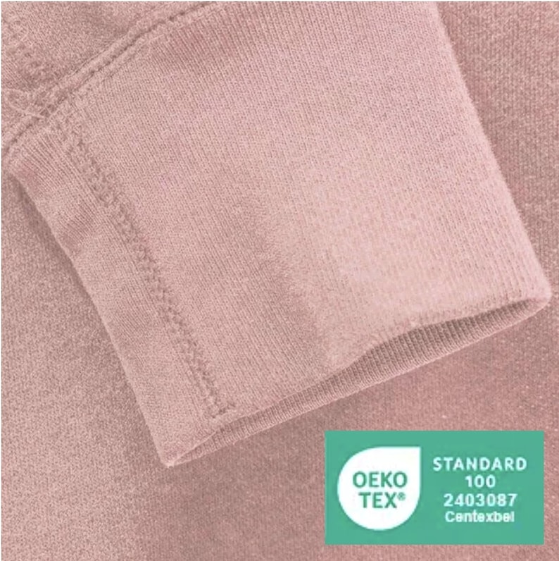 May include: Close-up of a dusty rose colored sweatshirt sleeve with a sewn seam. The sleeve has a green Oeko-Tex Standard 100 certification label with the number 2403087 and the text "Centexbel".