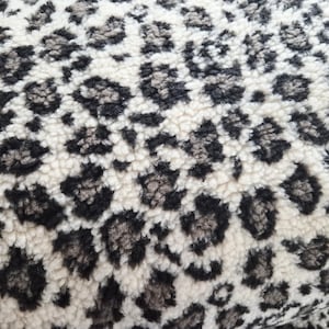 Könnte beinhalten: Nahaufnahme eines strukturierten Stoffes mit Leopardenmuster. Das Design zeigt schwarze und graue Flecken auf cremefarbenem Hintergrund. Der Stoff hat ein weiches, plüschiges Aussehen.