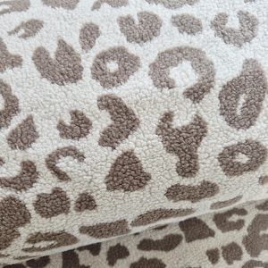 Könnte beinhalten: Nahaufnahme eines weichen, strukturierten Stoffes mit einem sich wiederholenden Leopardenmuster. Das Design zeigt braune Flecken auf cremefarbenem Hintergrund. Der Stoff scheint gerollt oder gefaltet zu sein und zeigt die Kontinuität des Musters.