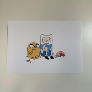 Può includere: Una stampa con i personaggi dei cartoni animati Finn e Jake di Adventure Time. Finn, con un cappello bianco e un completo blu, è seduto con Jake, un personaggio giallo simile a un cane. Sono raffigurati con cibo, su sfondo bianco.