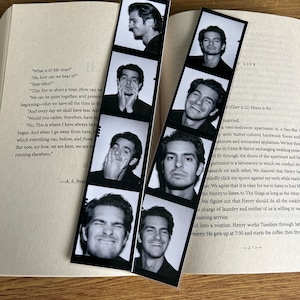 Peut inclure: Un marque-page en forme de bande photo noir et blanc avec le visage d'un homme dans chaque cadre. L'homme fait différentes expressions. Le marque-page est posé sur un livre avec du texte sur la vie d'un couple.