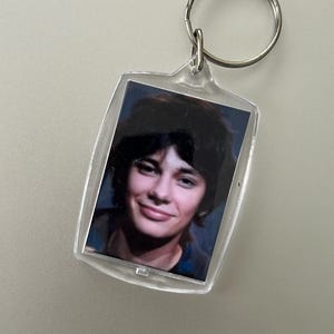 Può includere: Un portachiavi in plastica trasparente con una foto di un giovane con i capelli scuri e un sorriso. La foto è in una cornice rettangolare.