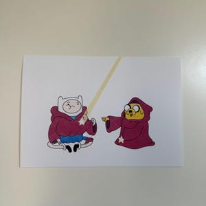 Può includere: Una stampa con i personaggi dei cartoni animati Finn e Jake di Adventure Time. Finn indossa un mantello con cappuccio bordeaux con una stella bianca e Jake indossa un mantello simile. Un raggio giallo si estende tra loro. Lo sfondo è bianco.