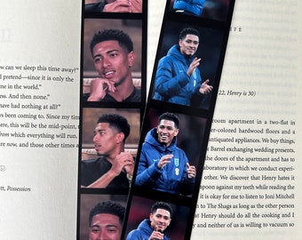 May include: Two photo strip bookmarks featuring images of a man in various poses. One strip shows the man in a black shirt, the other shows him in a blue jacket. The bookmarks are placed on an open book.