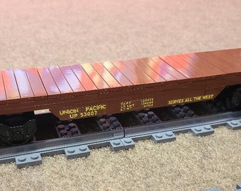 Op de afbeelding: Een bruine houten platte treinwagon met zwarte wielen en een zwart metalen frame. De wagon heeft een geel label met de tekst "UNION PACIFIC UP 53007" en "SERVES ALL THE WEST".