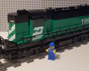 May include: A green and black Lego train engine with the number 1962 on the side. The engine has the words "Burlington Northern" printed on the side. A small Lego figure stands in front of the engine.