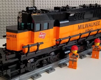Op de afbeelding: Een zwart en oranje modeltrein met de woorden "MILWAUKEE ROAD" op de zijkant. De modeltrein heeft het nummer "2071" op de zijkant. Twee miniatuur bouwvakkersfiguren staan voor de trein.