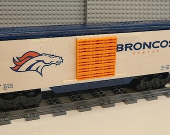 May include: A white Lego train car with a blue roof and orange details. The car has a Denver Broncos logo and the words "Broncos Denver" on the side.