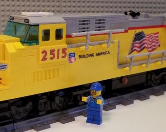May include: A yellow toy train engine with the number 2515 on the side. The engine has a black grill, a red stripe, and an American flag on the side. The engine is on a gray track and a small blue toy figure is standing in front of it.