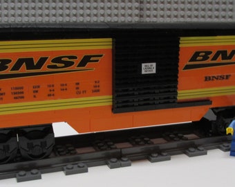 May include: A black and orange toy train car with the letters "BNSF" printed on the side. The car has a black roof and a black grate on the side. The car is on a gray track. A blue Lego figure stands next to the car.