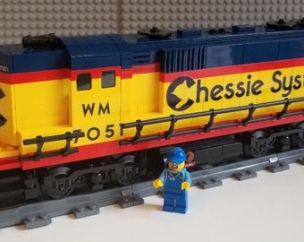 May include: A yellow, red, and blue toy train engine with the words "Chessie System" and "WM 7051" printed on the side. The engine is on a gray track and a small blue toy figure stands in front of it.
