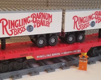 May include: A red toy train car with two white toy boxcars on top. The boxcars have the words "Ringling Bros. and Barnum & Bailey" printed on them. The train car has the words "Ringling Bros. and Barnum & Bailey" printed on the side. A small toy construction worker figure stands in front of the train.