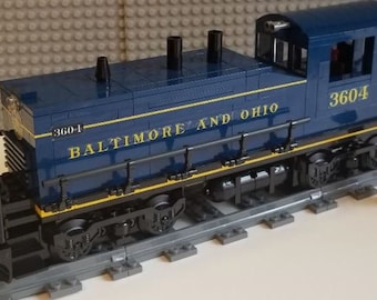 Op de afbeelding: Een blauwe Lego-treinlocomotief met de woorden "Baltimore and Ohio" en het nummer "3604" erop gedrukt. De locomotief staat op een spoor met een Lego-figuur ernaast.