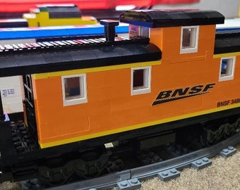 May include: A toy orange and black caboose with a black roof and a small black chimney. The caboose has a white window on each side and a BNSF logo on the side. The caboose is on a gray track.