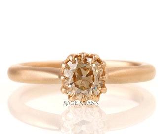 May include: A rose gold ring with a square-shaped, light brown gemstone set in a delicate, scalloped halo.