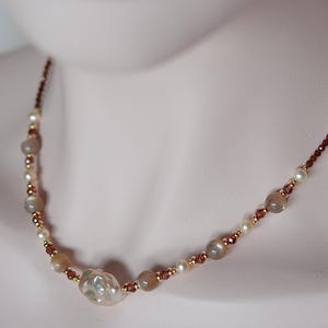 May include: An elegant necklace with a large, irregular pearl pendant, small pearls, moonstone beads, and tiny red-brown beads. The necklace is strung on a fine chain, creating a refined accessory. The necklace is suitable for special occasions.