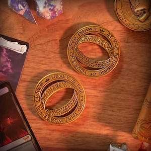 Puede incluir: Dos fichas Monarch doradas con texto grabado y diseño de corona, junto a cartas y dados sobre una superficie de madera. Las fichas presentan el texto "DRAW 1 AT THE BEGINNING OF YOUR END STEP" y "THE CROWN THAT WEARS THE HEAD IS HEAVY".