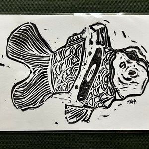 May include: Black and white linocut print of a goldfish with a detailed pattern on its scales. The fish is swimming to the right and has a large, round eye. The artist's initials "AMP" are signed in the bottom right corner.
