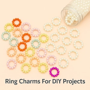 May include: A collection of colorful ring charms in various shades, including orange, pink, yellow, and green, scattered on a light peach surface. A clear container spills white ring charms. Text at the bottom reads "Ring Charms For DIY Projects."