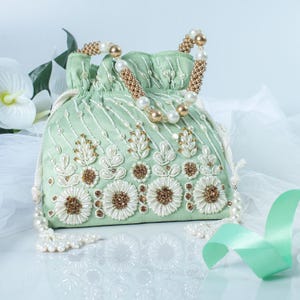 May include: A mint green drawstring bag with intricate floral embroidery in white and gold beads. The bag features a beaded handle with pearls and gold accents. The design includes white tassels and a mint green ribbon.