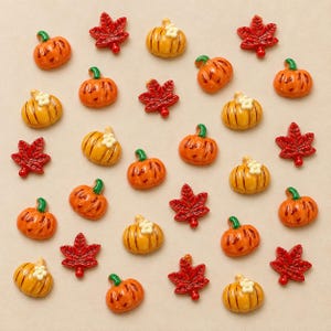 May include: A collection of small, decorative resin embellishments featuring orange and yellow pumpkins with green stems and red maple leaves. Some pumpkins are topped with small white flowers. These items are arranged in a scattered pattern.