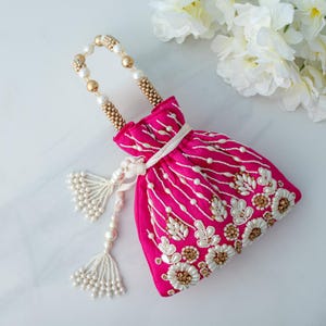 May include: A pink beaded drawstring bag with a gold beaded handle and white pearl tassels. The bag is decorated with white pearl and gold embellishments in a floral pattern.