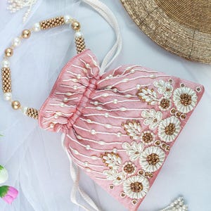 May include: A pink drawstring bag with a white pearl and gold bead embellishment. The bag has a floral design and a long white tassel.