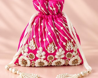 May include: A bright fuchsia drawstring pouch featuring intricate white beadwork and gold-toned floral embellishments. The bag is cinched at the top with white braided cords that end in decorative tassels made of white pearls and beads.