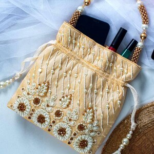 May include: A cream-colored drawstring bag with a beaded handle and floral embroidery. The bag is decorated with white and gold beads and has a tassel on the drawstring.