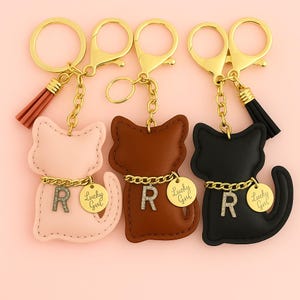 May include: Three cat-shaped keychains in pink, brown, and black. Each keychain has a gold-colored chain, a charm with the letter "R", and a "Lucky Girl" tag. The keychains have gold-colored key rings and tassels.