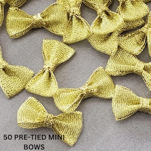 May include: A close-up of 50 gold pre-tied mini bows. The bows are made of a shimmery fabric and are perfect for adding a touch of sparkle to any project.