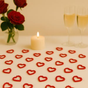 May include: A romantic scene featuring red heart-shaped decorations scattered on a white surface. A lit candle, two champagne flutes, and a vase of red roses complete the setting, creating a festive and loving atmosphere.
