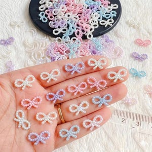 May include: A collection of pastel-colored bow-shaped beads. The beads are in shades of pink, purple, blue, and white. Some of the beads are held in a hand, while others are scattered around a black surface. A ruler is visible in the image.