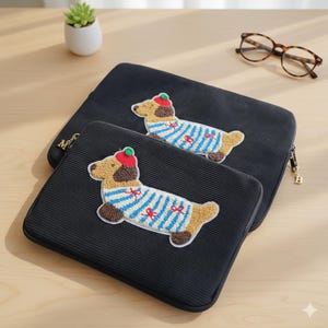 May include: Two black laptop sleeves with a corduroy texture. Each sleeve features an embroidered dachshund wearing a blue and white striped sweater and a red hat. A small succulent plant and eyeglasses are also visible.